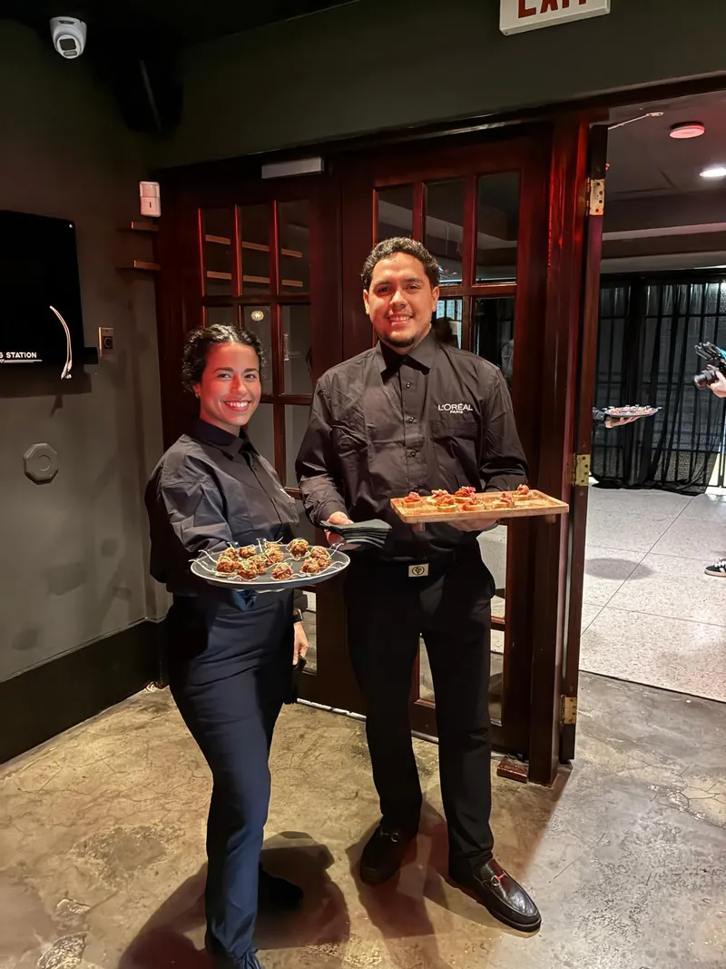 Dos personas del equipo, de uniforme negro, con bandejas de canapés y croquetas listas para pasar entre los invitados a la entrada del salón.